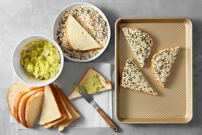 overhead shot; light grey background; A bowl of creamy shrimp mixture; Slices of white bread are ready to be topped with the mixture and coated in sesame seeds; A baking sheet lined with parchment paper awaits the assembled toasts