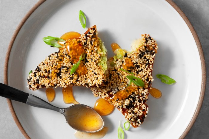 overhead shot; light grey background; A plate of crispy shrimp toast is presented, showcasing a golden brown exterior and a sesame seed coating; The toast is cut in half with some drizzle of sweet chili sauce, a silver spoon is also placed in the plate