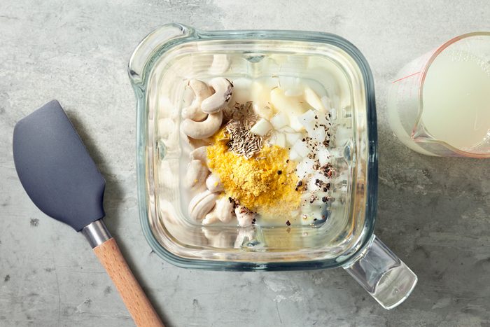 Overhead shot of drain and rinse cashews; place soaked cashews; pasta water; onion; garlic; nutritional yeast; lemon juice; salt; pepper and rosemary in a blender; spatula; grey marble surface