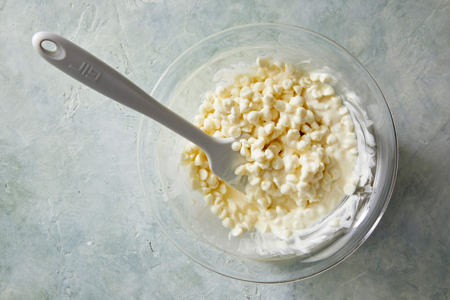 overhead shot; white background; A glass bowl contains a mixture of melted white chocolate chips; A spatula is resting in the bowl