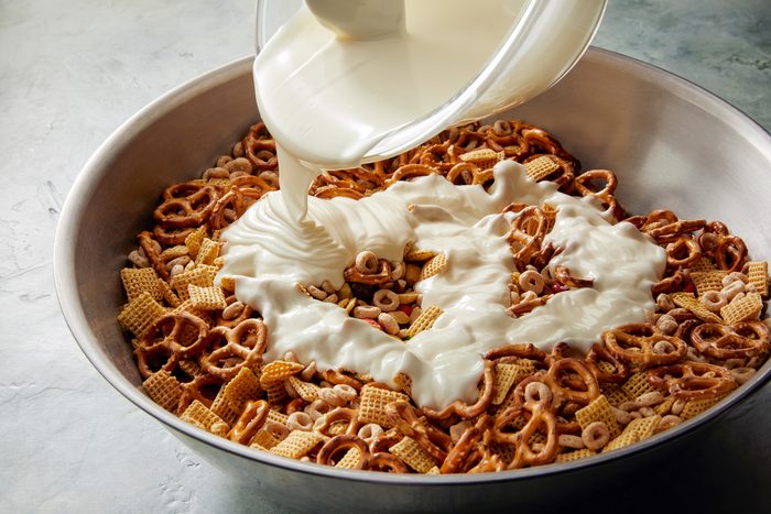 3/4th shot; white background; a bowl of mixed nuts and pretzels being coated with melted white chocolate; The chocolate is being poured from a pitcher onto the mixture