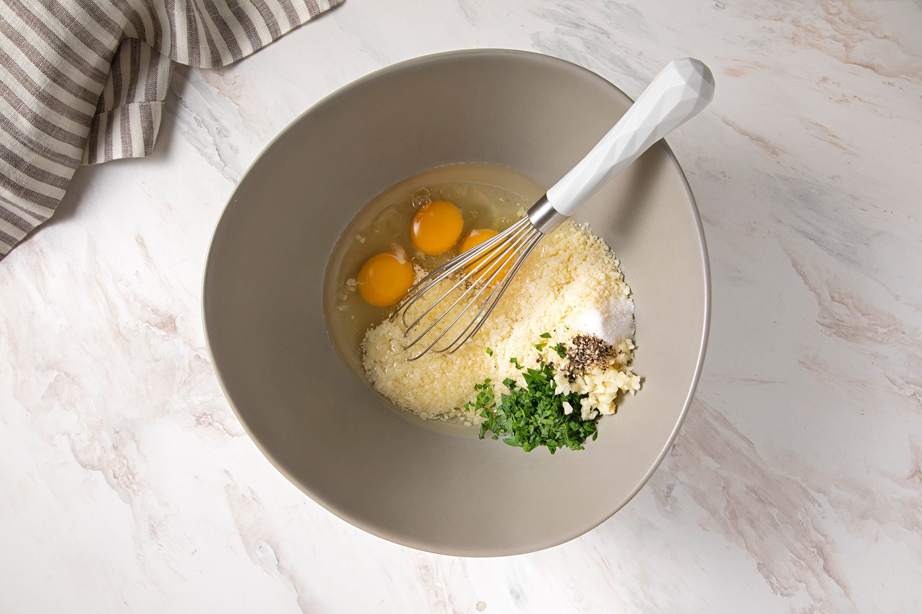 Top-down view of a mixing bowl containing three cracked eggs, grated Parmesan cheese, chopped garlic, chopped parsley, and black pepper. A whisk with a white handle rests inside the bowl. A striped cloth is partially visible in the top left corner.