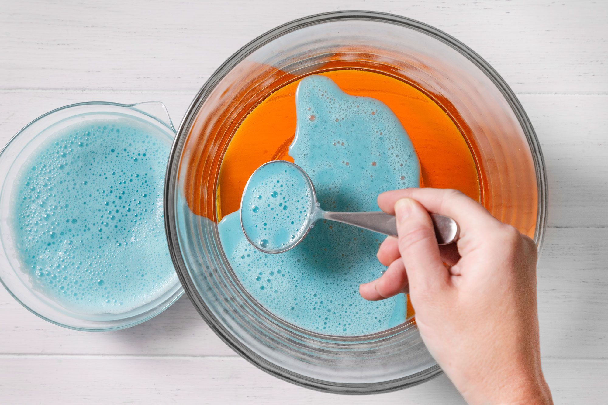 A person is holding a spoon in their right hand over a glass bowl, the bowl contains an orange liquid that is bubbling and foam on top of the liquid
