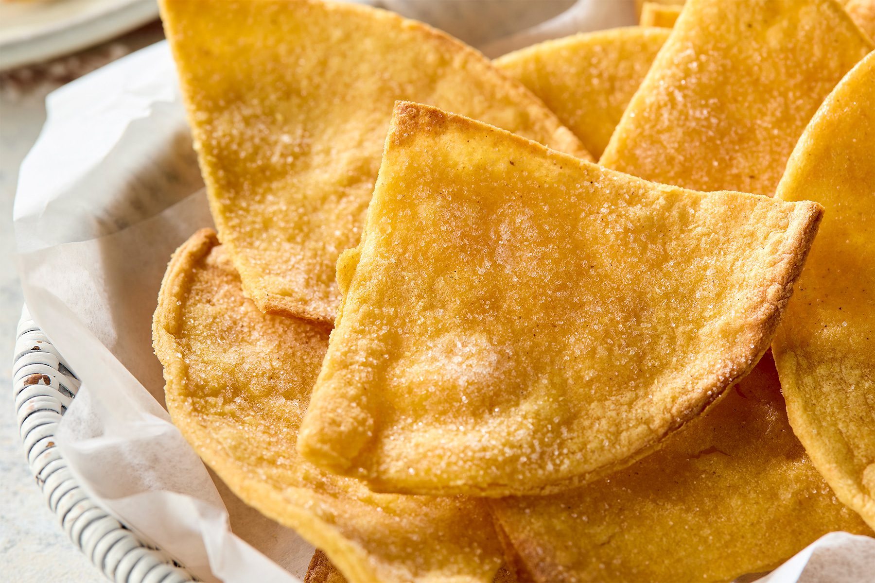 a close-up of a pile of golden brown tortilla chips, The chips are covered in a light dusting of salt, which adds a layer of texture and flavor, The chips are arranged in a basket lined with white parchment paper;