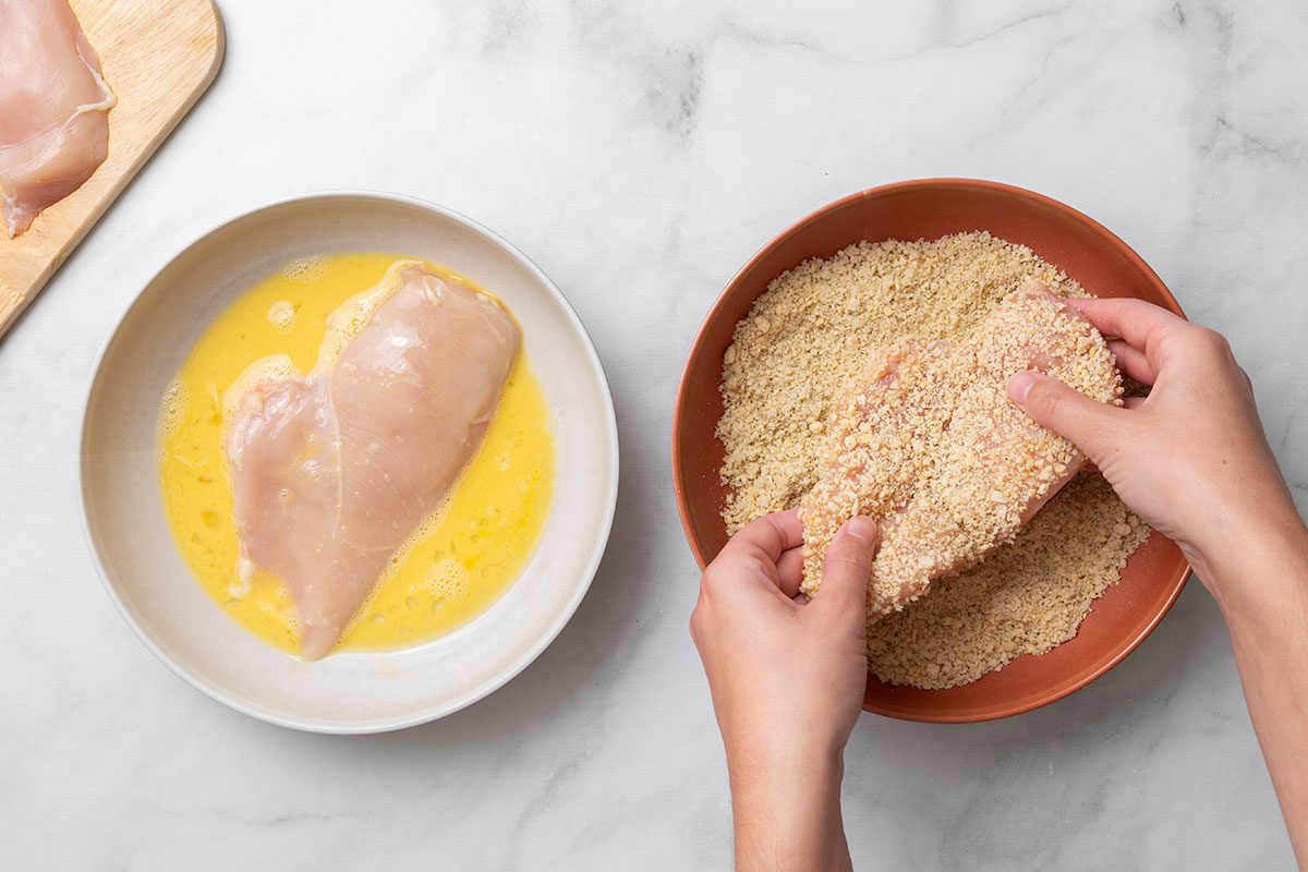 A chicken breast dipped in a bowl with a beaten egg and another chicken breast being coated in seasoned panko breadcrumbs in a seperate bowl for step one of Baked Chicken Parmesan recipe for Taste of Home