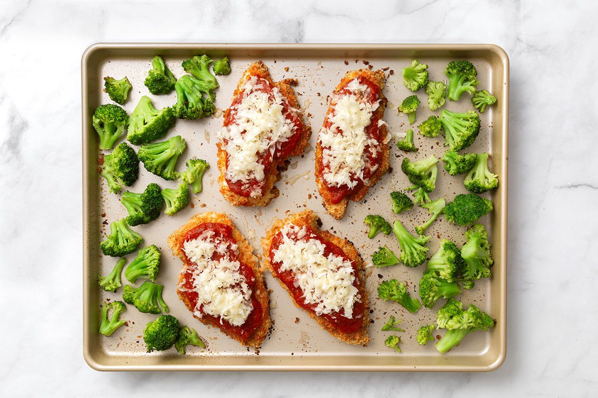 Baked chicken breasts topped with marinara sauce and shredded mozzarella cheese on a baking sheet with roasted broccoli florets for step three of Baked Chicken Parmesan recipe for Taste of Home