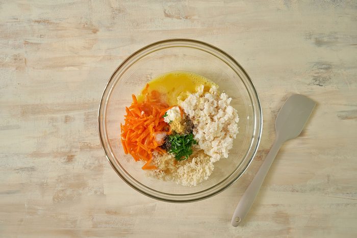 Overhead view of crabmeat, bread crumbs, shredded carrot, egg, melted butter, parsley, mayonnaise, Worcestershire sauce, mustard, salt, and pepper being gently folded together in a large bowl for the Taste of Home Baked Crab Cakes recipe.