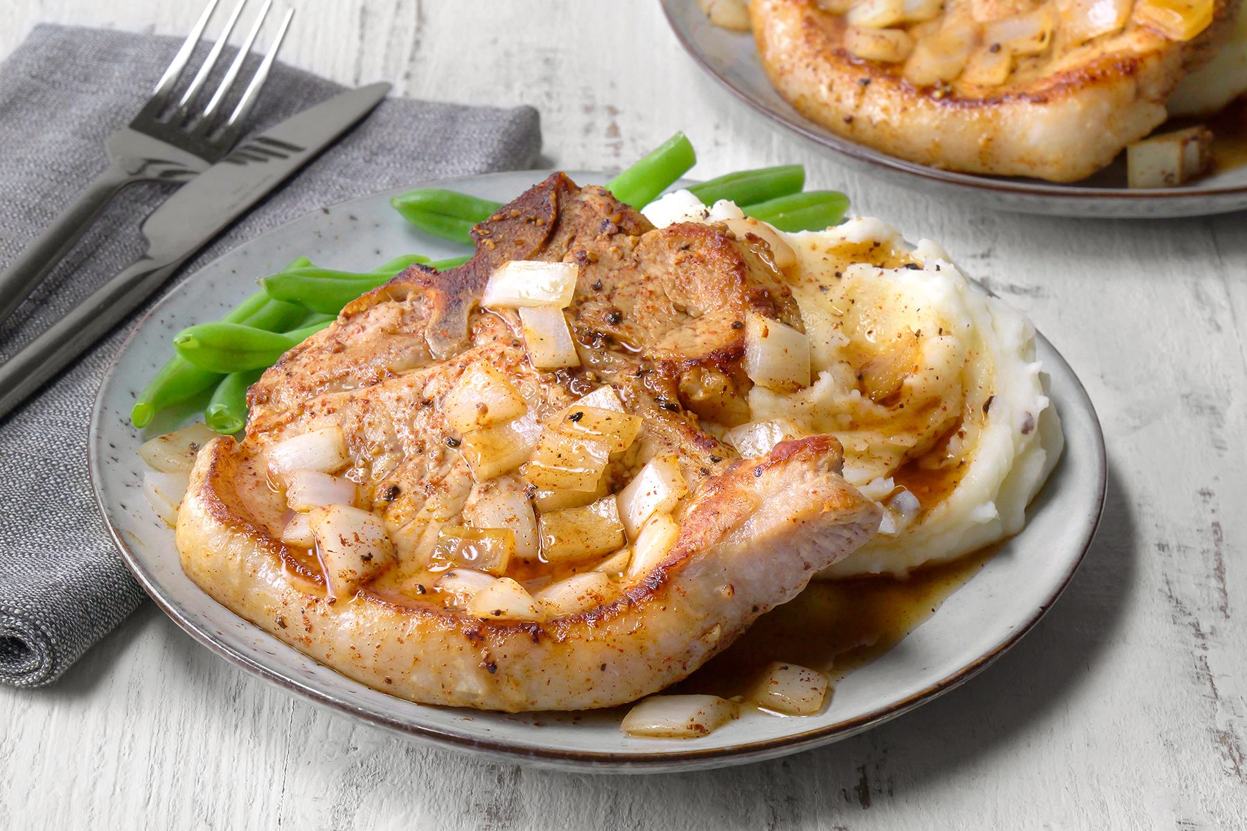 A plate with a cooked pork chop topped with onions, served with mashed potatoes and green beans. A fork and knife are placed on a grey napkin beside the plate, all set on a light wooden surface.