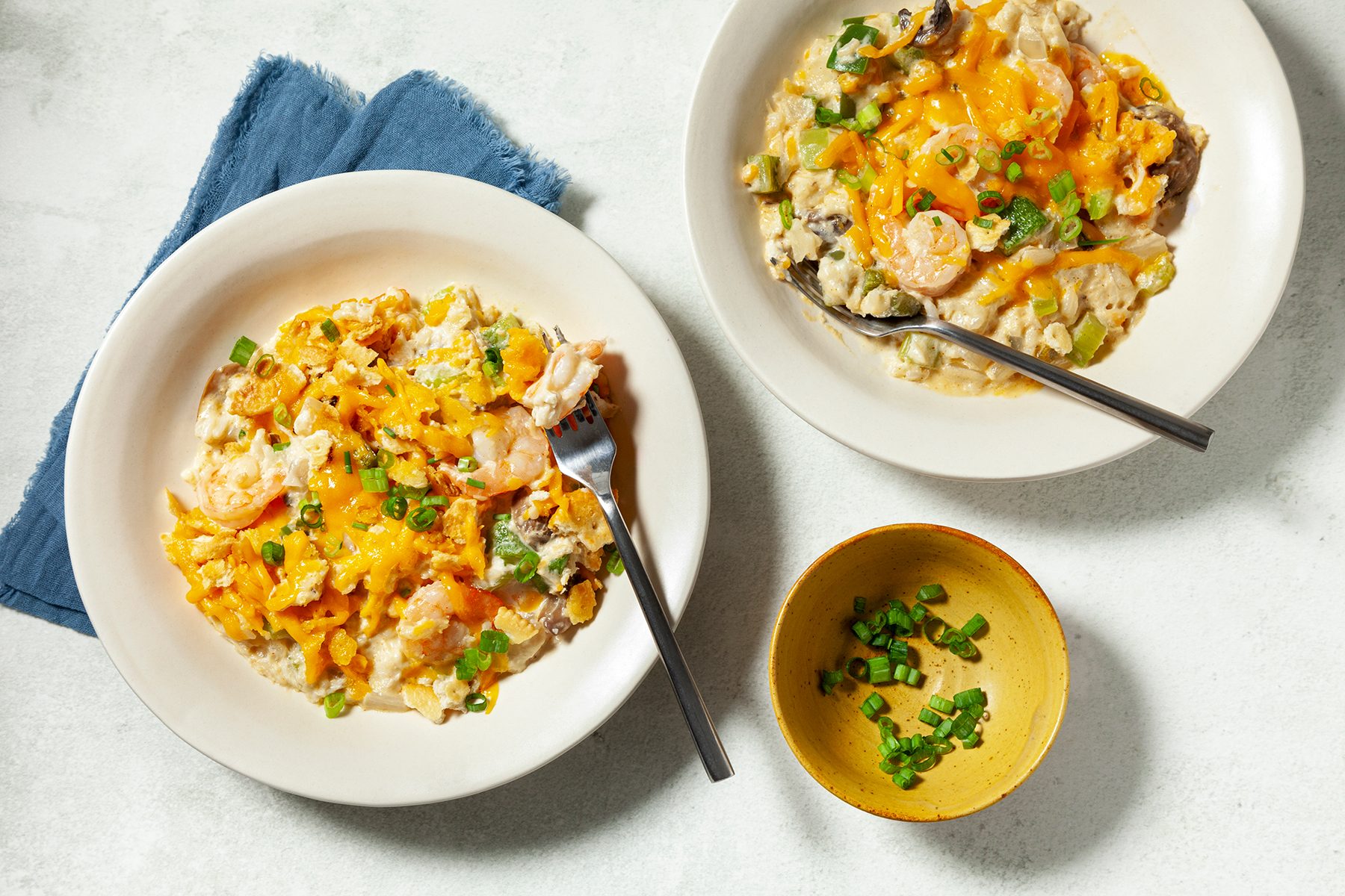 Two dishes of shrimp and vegetable casserole topped with melted cheese and chopped green onions, accompanied by a small yellow bowl of additional green onions. A dark blue napkin sits beside one plate on a light surface.
