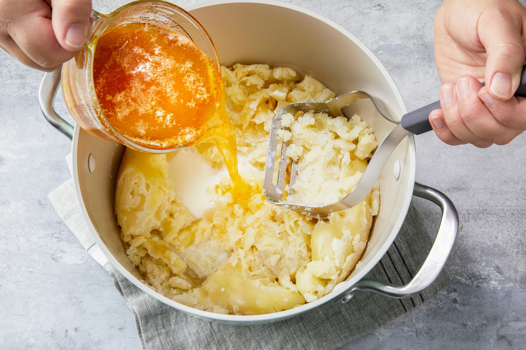 A bowl of cooked potatoes being gently mashed with a potato masher. Milk and a mixture of browned butter with garlic are being poured in gradually, with a creamy texture forming as the ingredients combine.