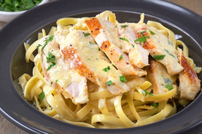 3/4th shot of a bowl of Chicken Alfredo is showcased, featuring tender pieces of grilled chicken nestled in a creamy, buttery Alfredo sauce, Fettuccine pasta forms the base of the dish;