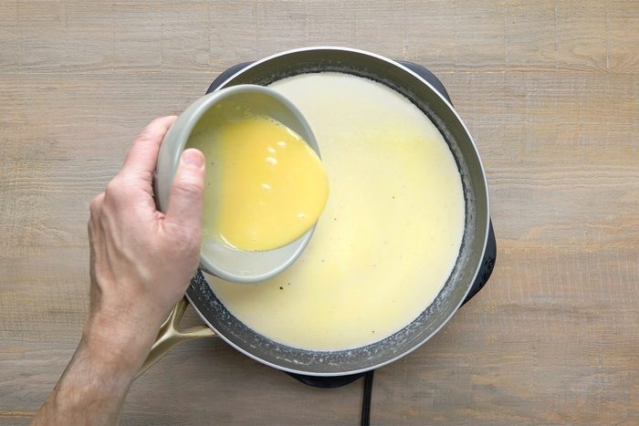 overhead shot of a person pouring a bowl of whisked egg yolks into a skillet filled with a creamy, pale yellow sauce, The egg yolks are creating ripples in the sauce, The skillet is placed on a wooden surface;