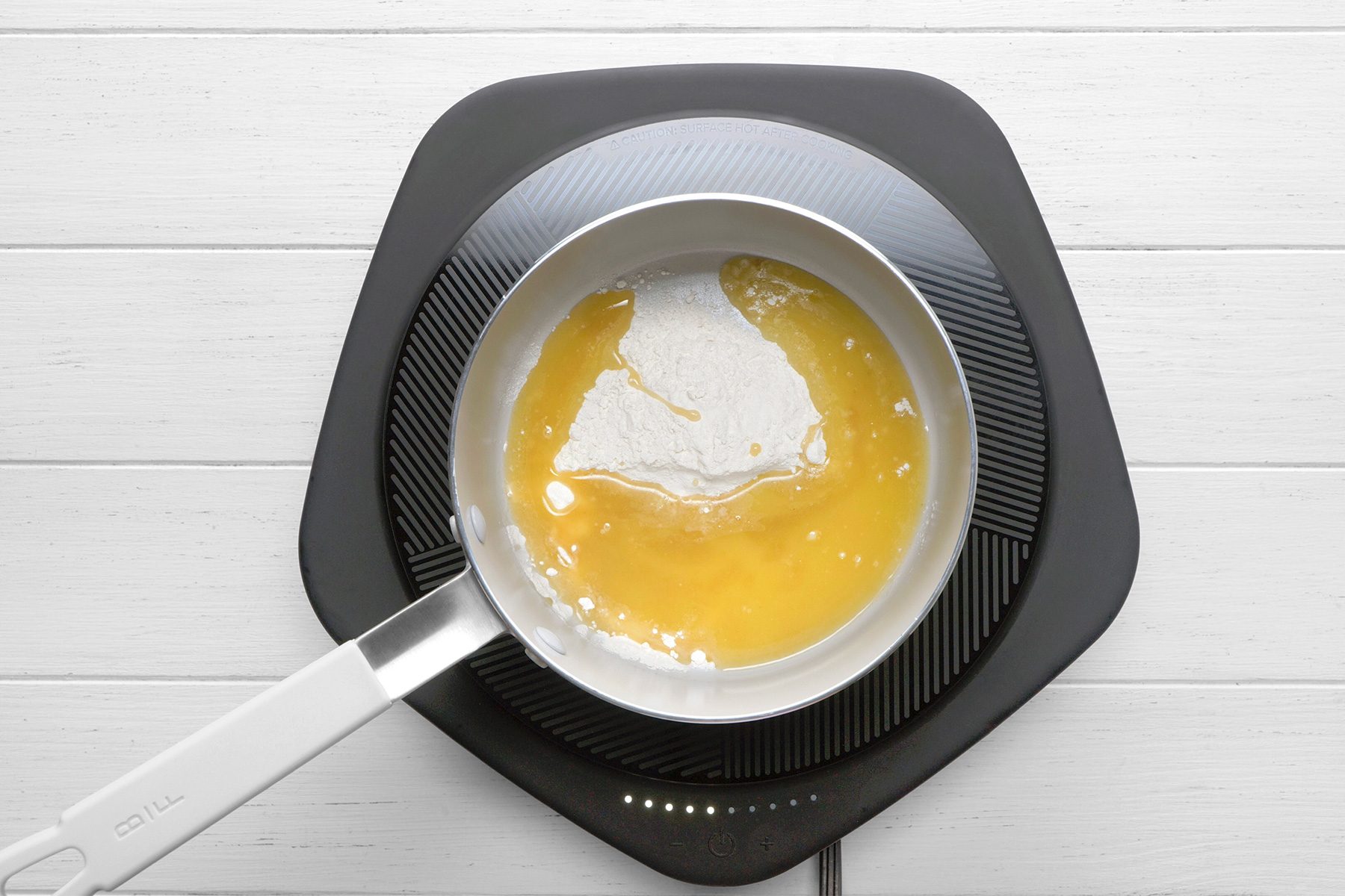 overhead shot of a small saucepan on an electric induction cooktop, The saucepan contains melted butter and a sprinkling of flour, The butter is a golden brown color, and the flour is a white powder, The mixture is starting to bubble and sizzle, indicating that it is heating up;
