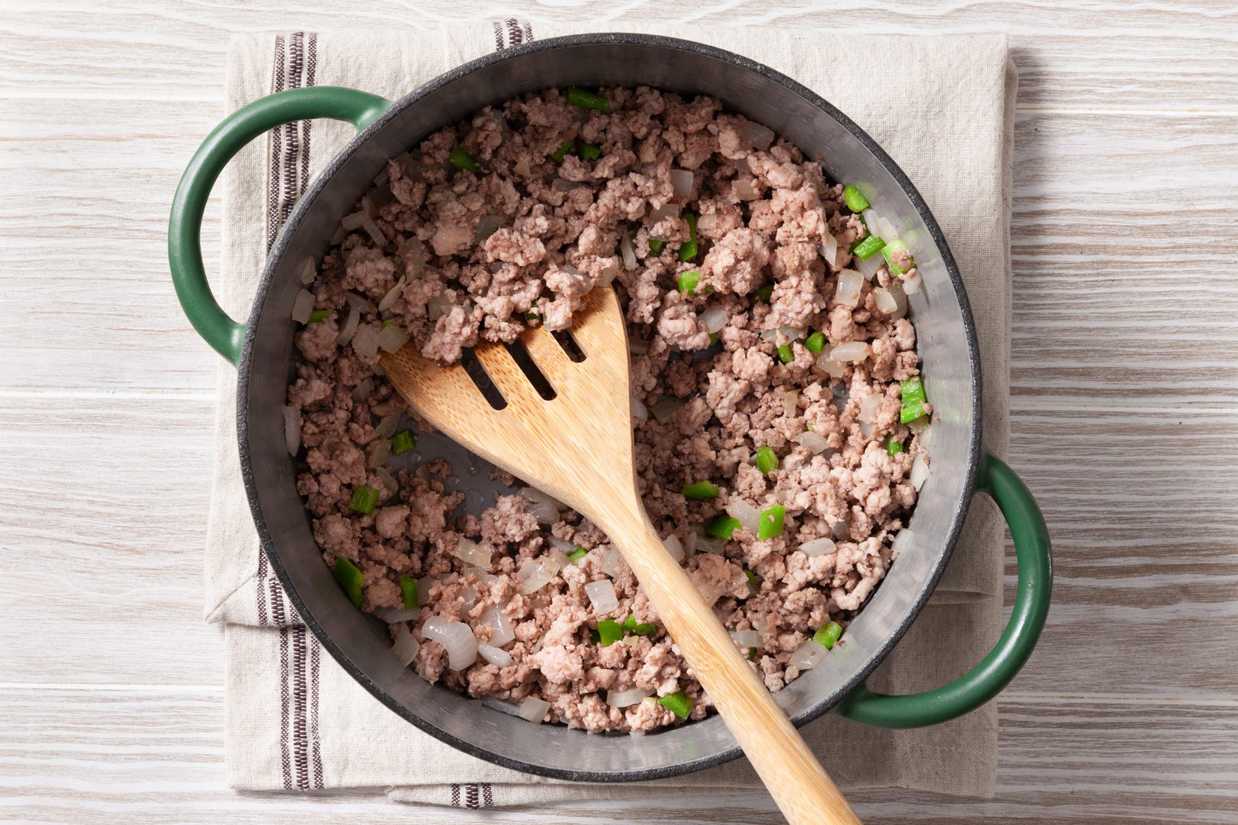 An overhead shot of a green cast-iron pot on a wooden surface. The pot contains cooked and crumbled ground meat, likely ground beef, mixed with chopped green onions. A wooden spatula rests in the pot, and a kitchen towel is placed underneath.