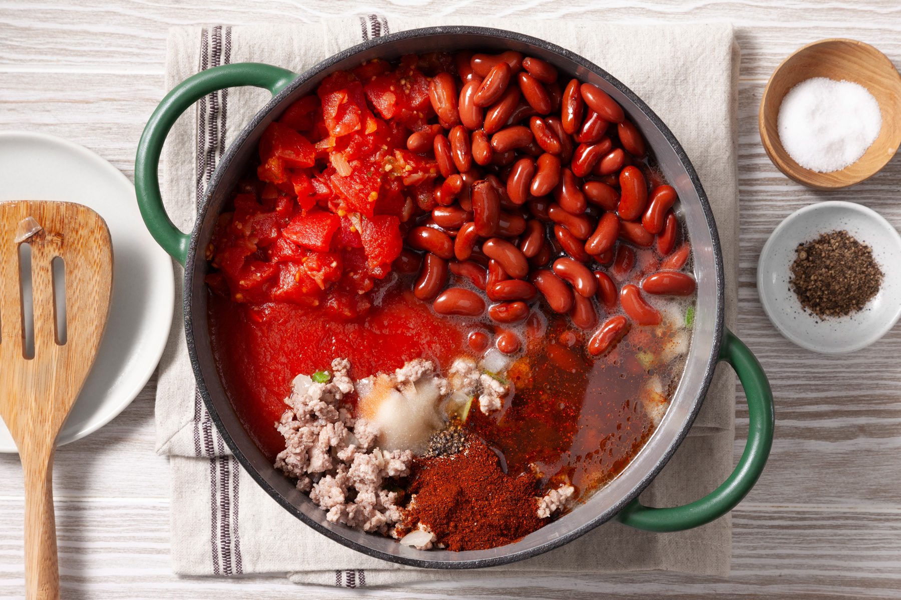 An overhead shot of a green cast-iron pot filled with various ingredients for a chili recipe. Ground meat, diced tomatoes, kidney beans, and a liquid base are visible in the pot. A wooden spatula rests on the rim, and a small bowl of salt and a bowl of ground black pepper are placed nearby.