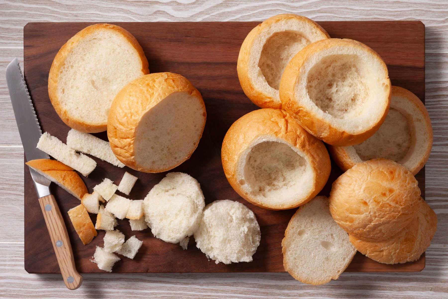 An overhead shot of several bread rolls arranged on a wooden cutting board. Some of the rolls have been hollowed out, creating bread bowls, while others have been cut into small cubes. A knife rests on the board, likely used to hollow out the rolls and cut them into cubes.