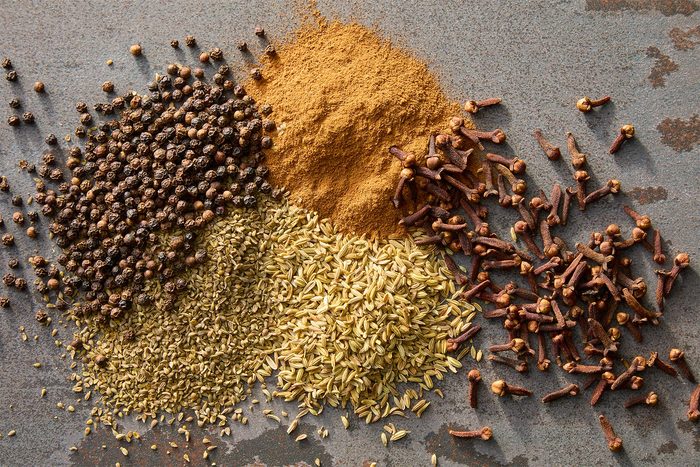 A flat lay of assorted spices on a gray surface, featuring a pile of black peppercorns, ground cinnamon, fennel seeds, and whole cloves, arranged in a visually appealing manner.