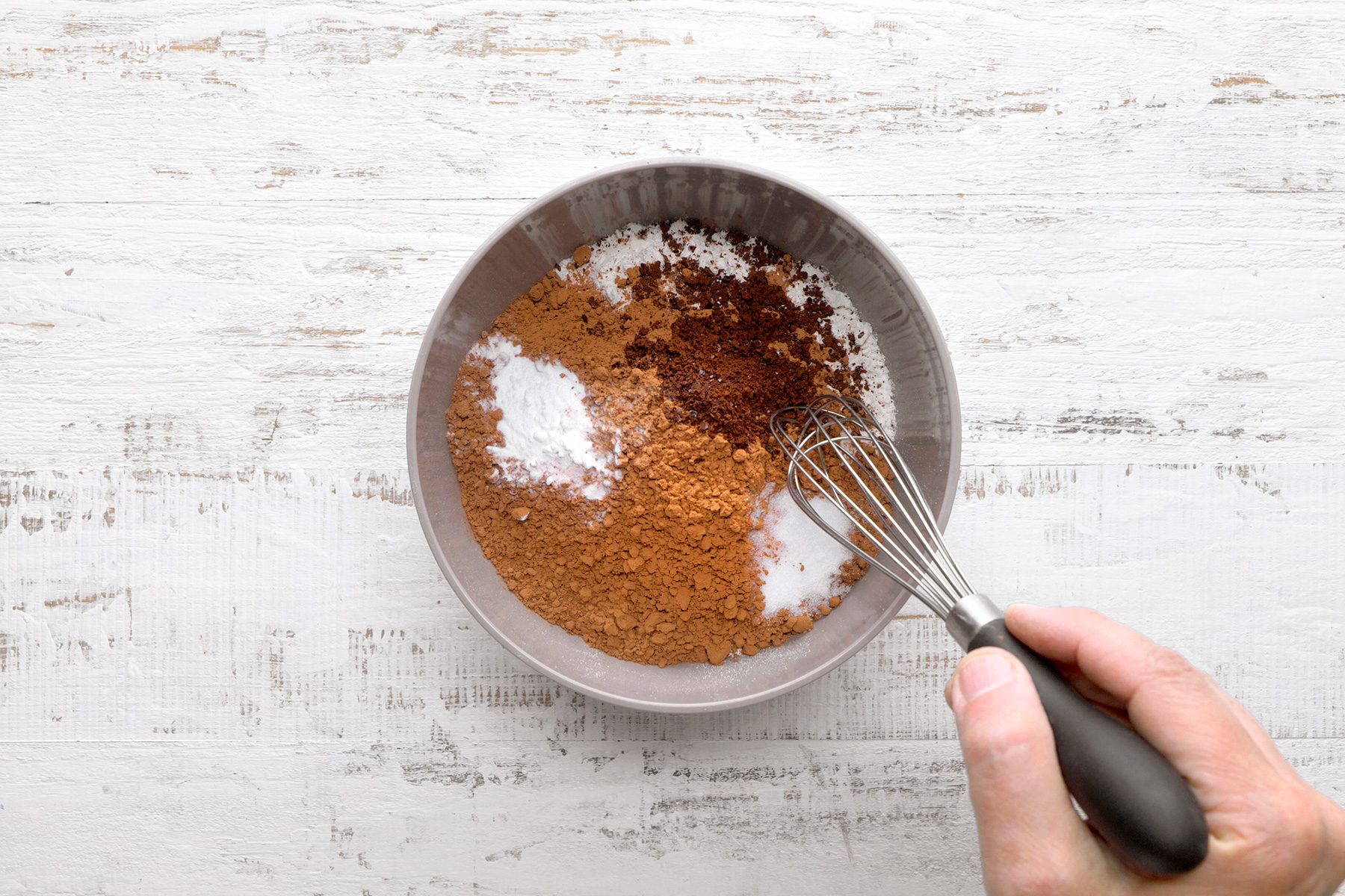 A hand holds a whisk above a bowl containing cocoa powder, baking soda, and sugar on a white wooden surface. The ingredients appear ready to be mixed.