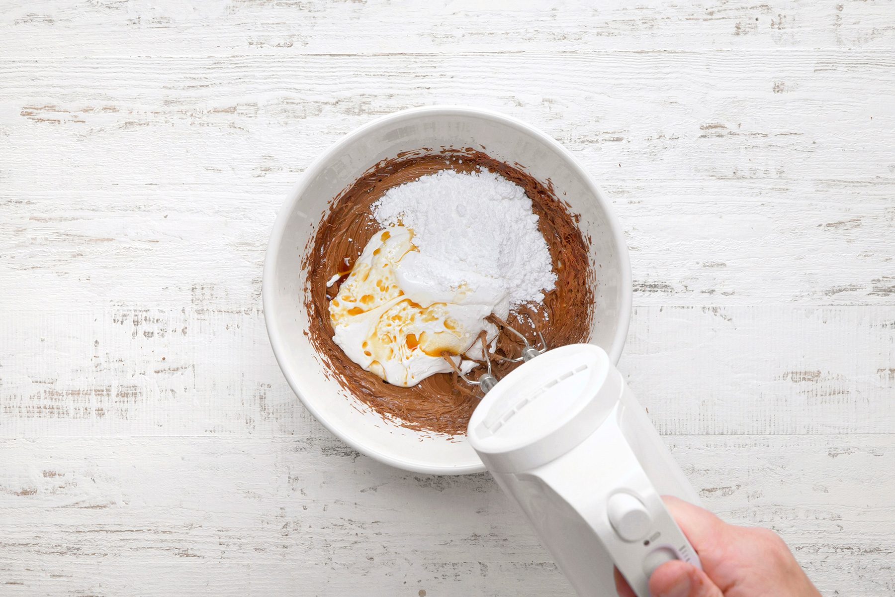 A white hand mixer is partially submerged in a bowl containing chocolate mix, cream cheese, and vanilla extract on a white wooden surface.