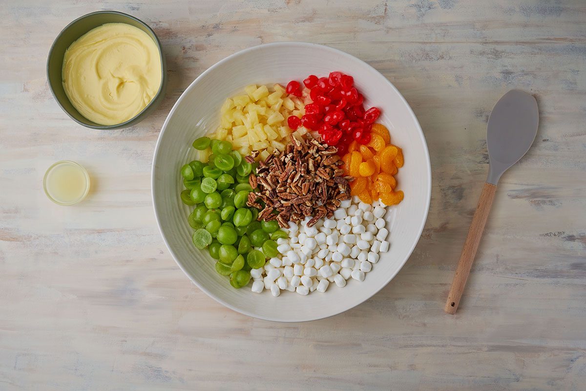 TA colorful arrangement of fruit salad ingredients mixed together including grapes, marshmallows, mandarin oranges, and pecans in a large bowl.