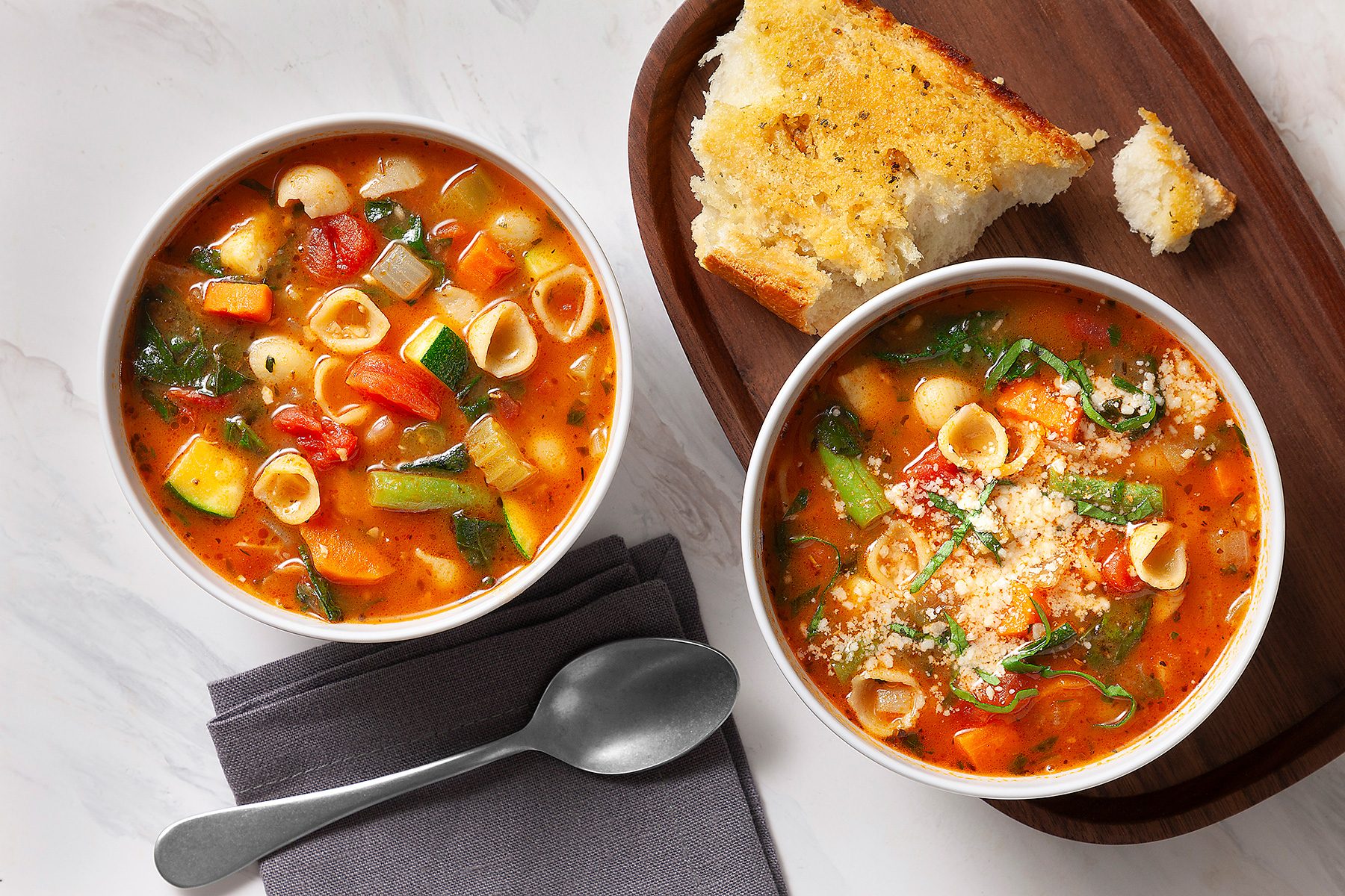 Two bowls of vegetable soup with shell pasta and greens, topped with grated cheese. A slice of crusty bread on a wooden board sits beside the soup. A spoon rests on a folded napkin on a marble surface.