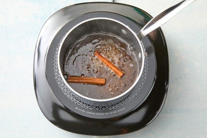 A pot on a black stovetop contains a dark liquid with two cinnamon sticks floating in it. A metal ladle rests on the side of the pot.