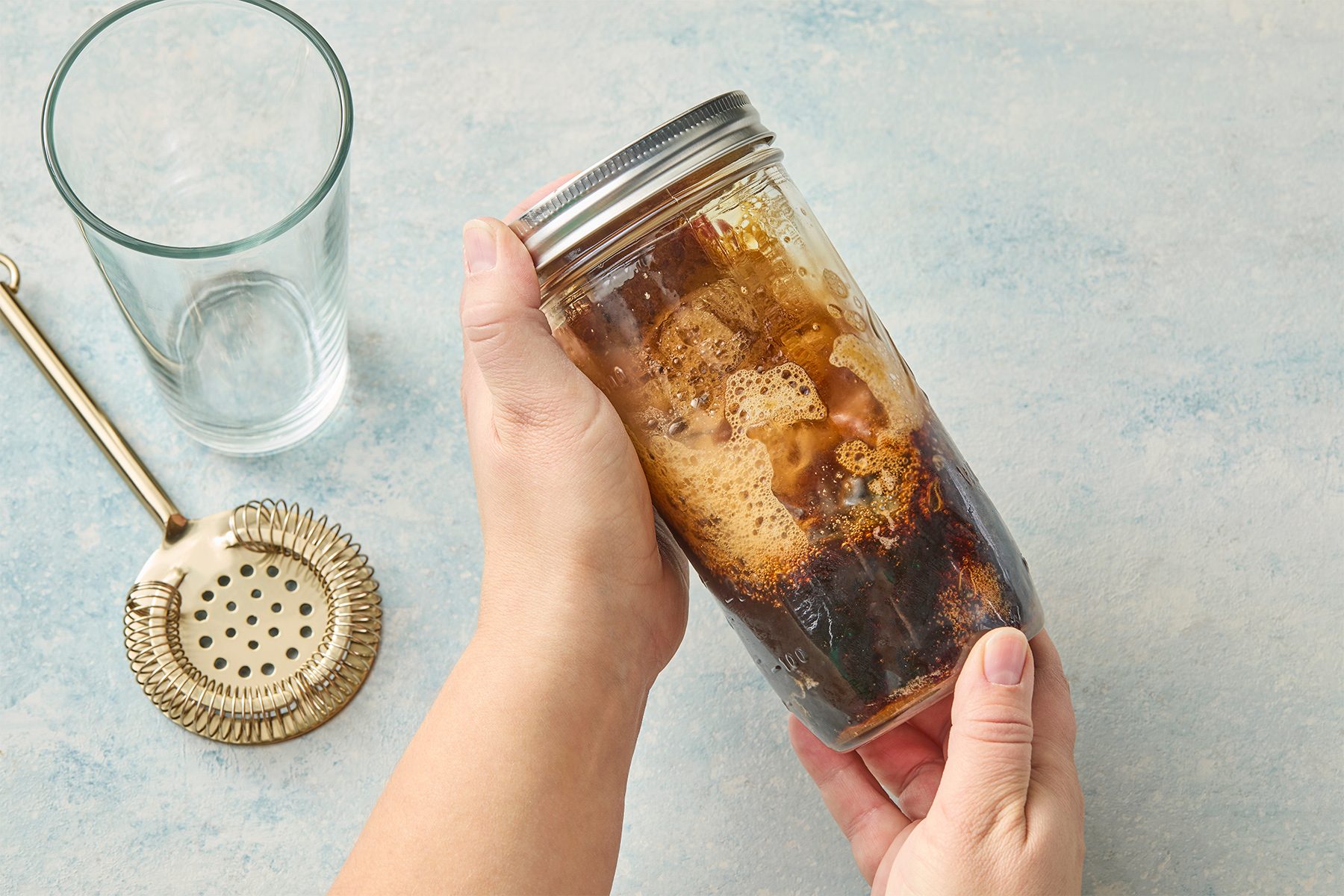 A person holding a glass jar filled with a foamy brown liquid. An empty glass, a strainer, and a spoon are on a light blue surface nearby.
