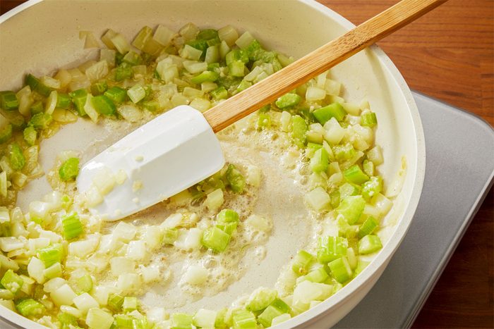 3/4th shot of a white pan filled with chopped onions and celery that are being sautéed in butter or oil, A white spatula is being used to stir the ingredients, The pan is placed on an electric induction cooktop