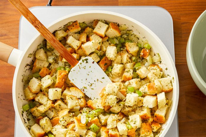 overhead shot of a white pan filled with a mixture of diced bread, chopped celery, and chopped onions, The bread cubes are a light brown color, and the vegetables are a mix of green and white, A wooden spatula is being used to stir the ingredients together