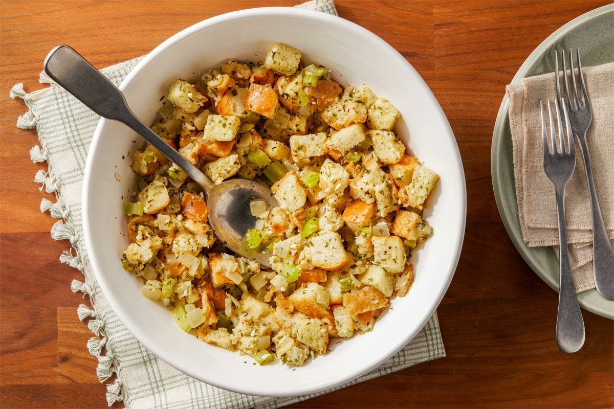 shot of a white bowl filled with a mixture of diced bread, chopped celery, and chopped onions, The bread cubes are a light brown color, and the vegetables are a mix of green and white, The mixture is seasoned with herbs and spices, bowl is placed over white kitchen cloth, A spoon is resting in the bowl;
