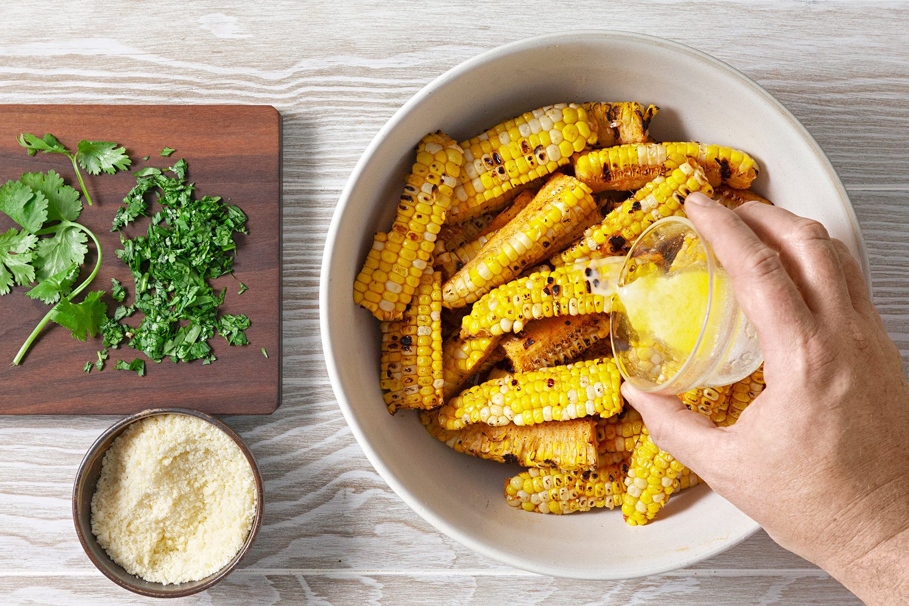 Overhead shot of place corn ribs in a large bowl; drizzle with melted butter and sprinkle with Parmesan cheese and cilantro; toss to coat; serve with lime wedges and additional Parmesan cheese; wooden surface;