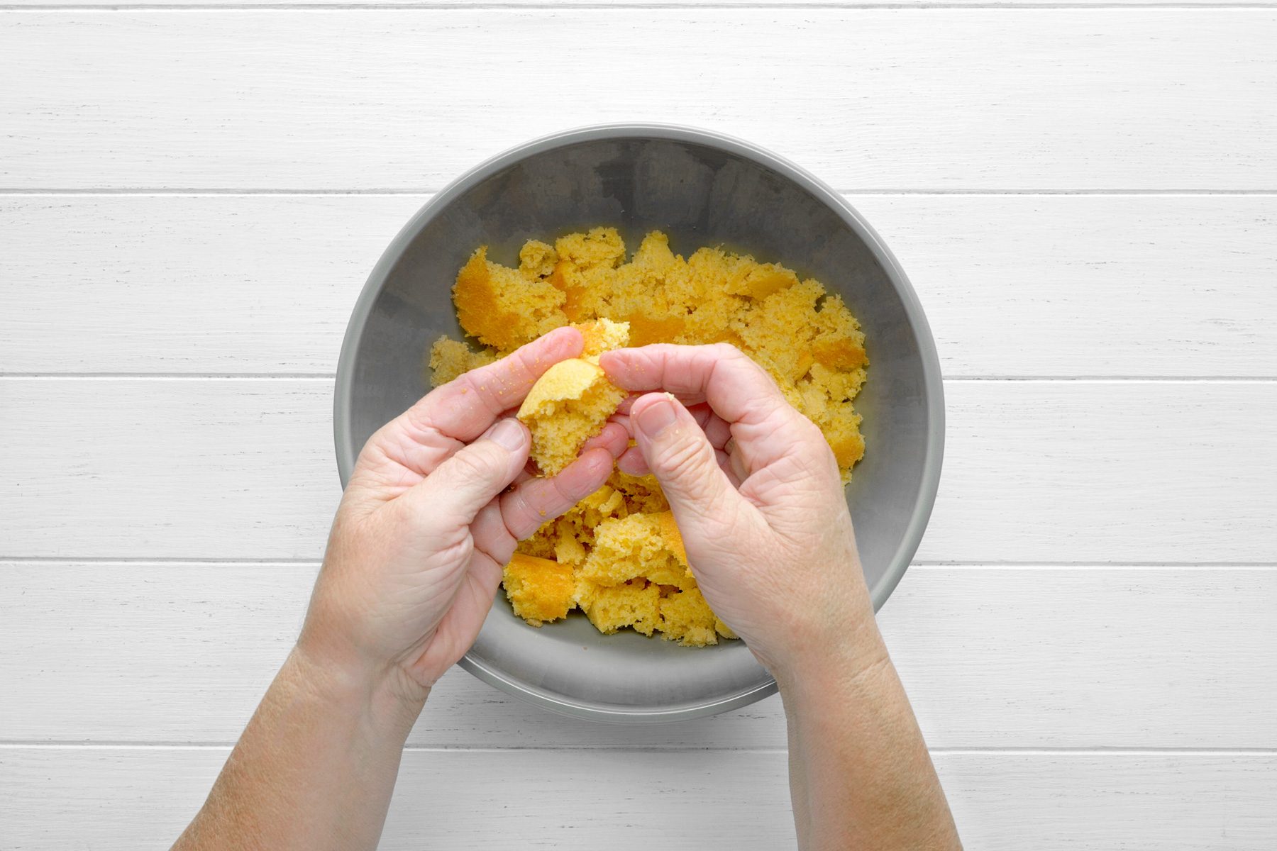 overhead shot of a hand tearing apart a piece of cornbread in a gray bowl, The cornbread appears to be golden brown and crumbly, The hand is breaking the cornbread into smaller pieces, The bowl is placed on a white wooden surface;