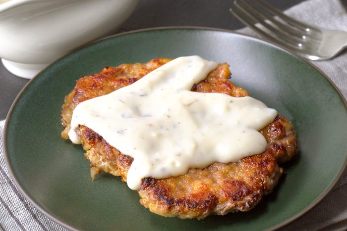 3/4th shot of a delicious Country Fried Steak served on a plate, A breaded and pan-fried steak patty is generously covered in a creamy white gravy, The steak has a golden brown exterior and appears tender and juicy;