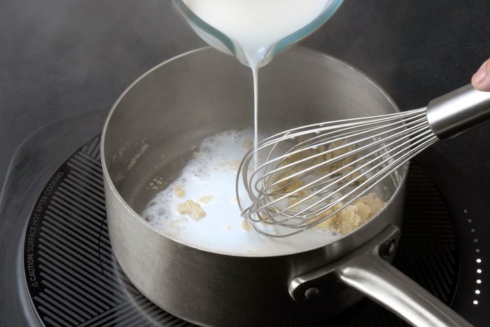3/4th shot of a hand pouring a liquid into a saucepan on an electric induction cooktop. The saucepan contains a mixture of melted butter and flour, and the liquid is being added gradually while whisking; The liquid appears to be milk or cream, and the mixture is starting to thicken;