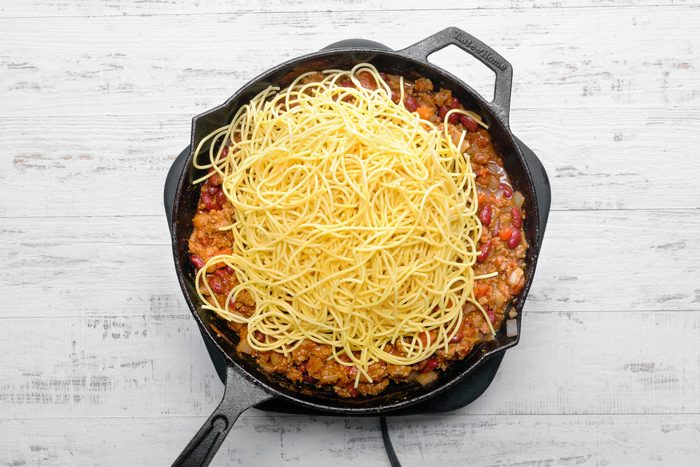 Drained spaghetti put into a skillet filled with bacon, beans, sauces and spices