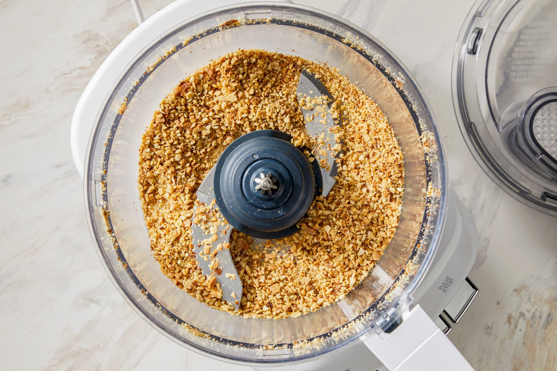 A food processor in action, with the lid on and almonds visibly chopped into fine pieces inside the bowl.