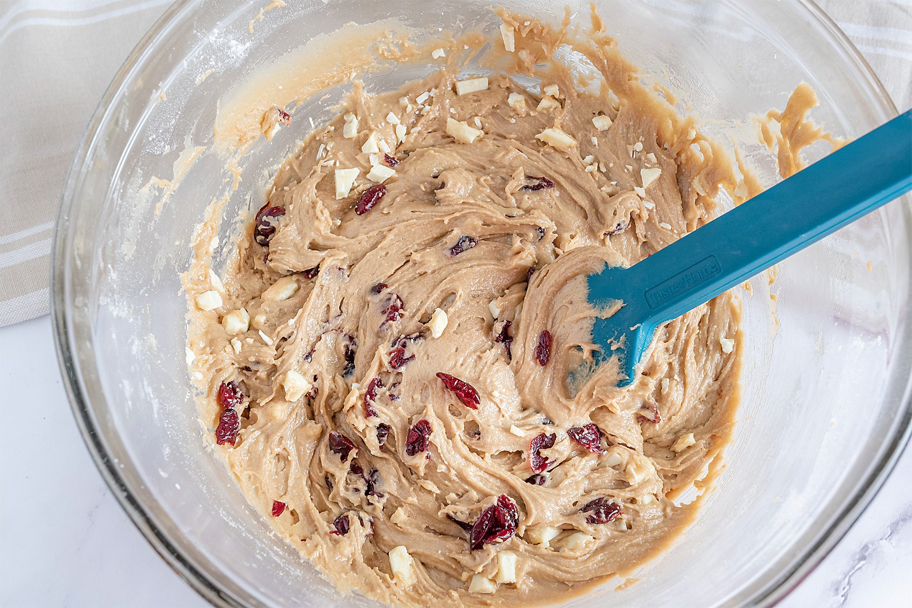 A glass bowl filled with cookie dough containing white chocolate chunks and dried cranberries. A blue spatula rests in the mixture, which is set on a white surface.