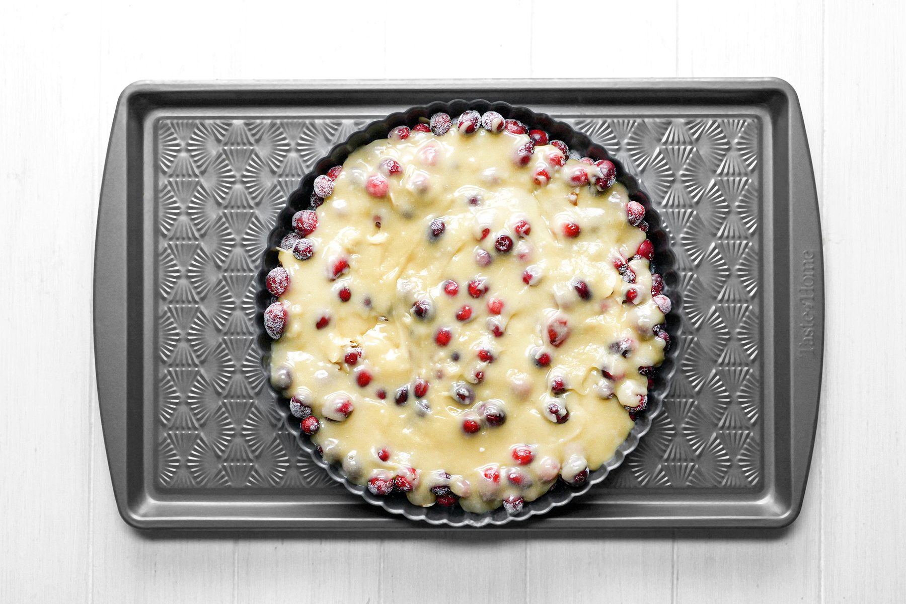 A round baking tin filled with unbaked cake batter and cranberries sits on a textured baking tray. The cranberries are partially submerged in the creamy batter. The background is a light wooden surface.