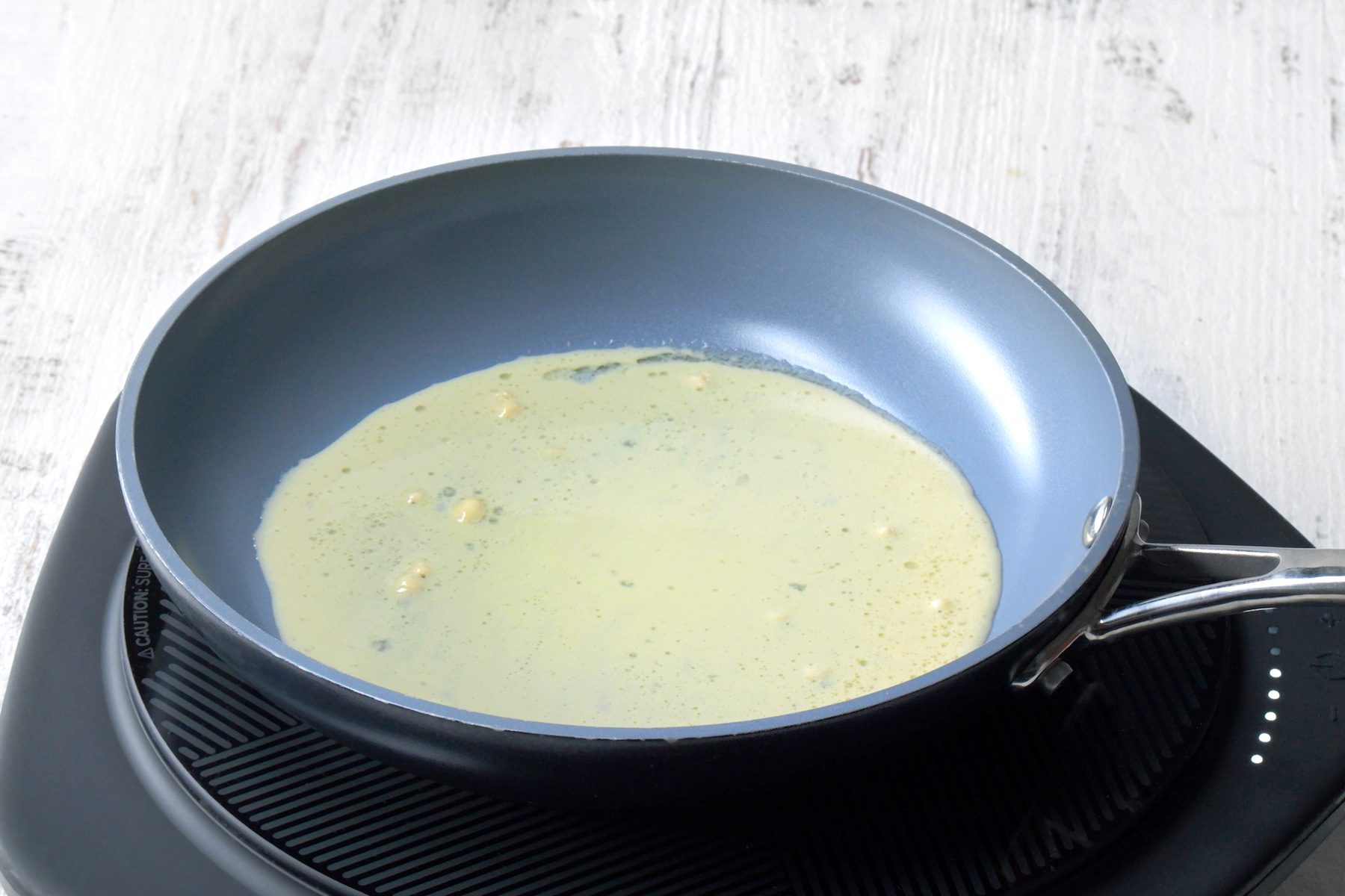 3/4th shot; white wooden background; a blue frying pan on a stove, Inside the pan, there’s a liquid mixture, possibly batter for pancakes or crepes, with small lumps