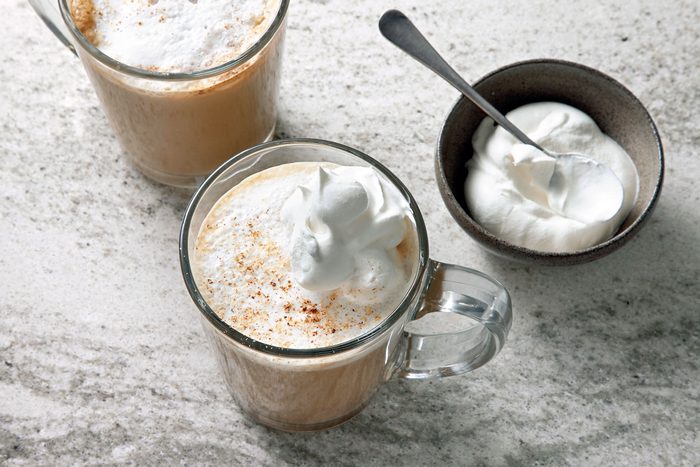 Two glasses of coffee, one with whipped cream and the other with a layer of cinnamon on top are sitting on a light grey surface