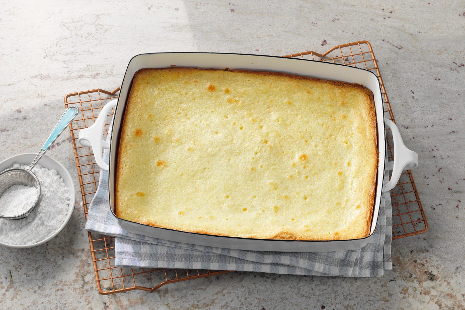 A golden-brown pudding in a white rectangular baking dish, placed on a checkered cloth and wire rack. A bowl of powdered sugar with a spoon is beside the dish on a light stone surface.