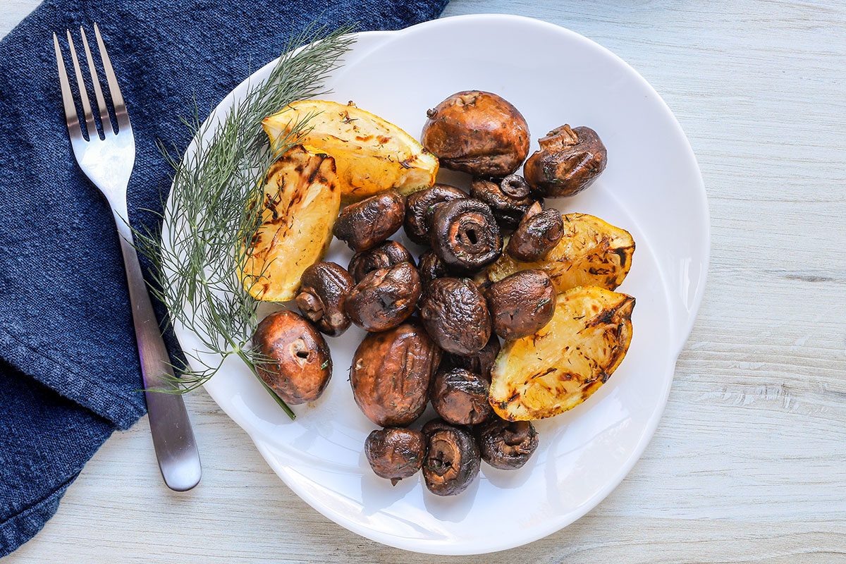 Grilled mushrooms on a plate with grilled lemon wedges and a sprig of dill.