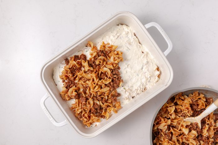 Hamburger noodle casserole being assembled in baking dish.