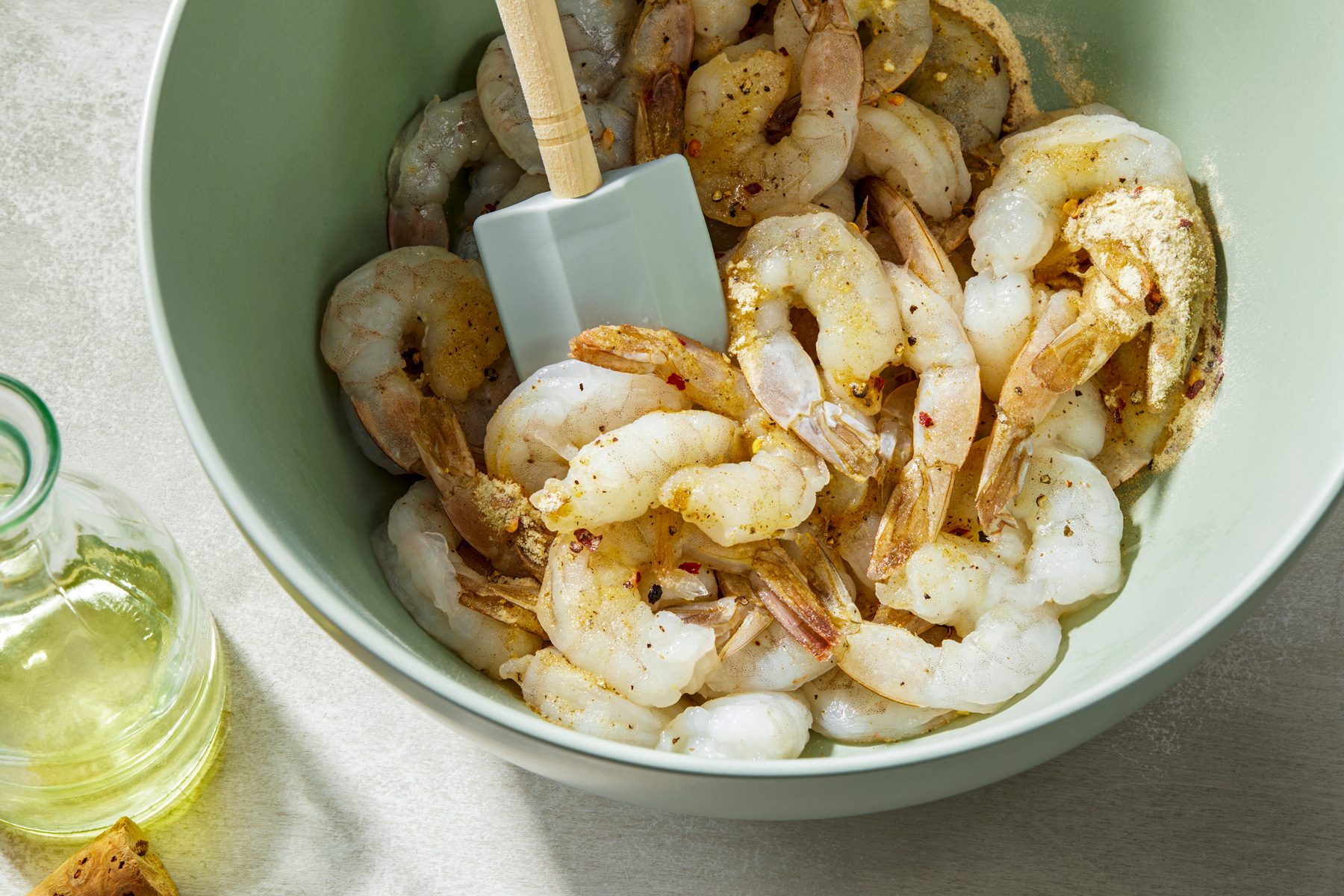 3/4 shot of a green bowl filled with raw shrimp coated in spices, with a spatula resting inside and a bottle of olive oil visible in the background.