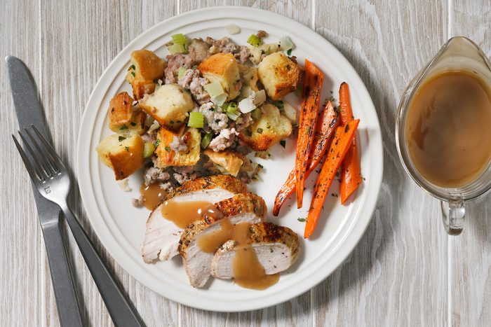 Overhead shot of a plate of food on a white wooden table; the plate has a white background and a white rim and it contains a variety of foods including Sausage Stuffing and carrots; there is also a gravy boat to the right of the plate which is filled with gravy; a knife and fork placed beside the table.