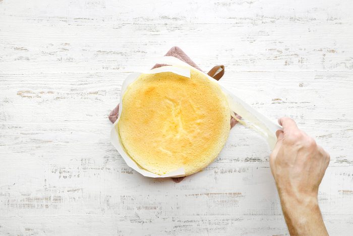 overhead shot of a hand peeling back a piece of parchment paper from a baked cake, The cake is a light golden brown color and has a domed shape, The parchment paper is sticking slightly to the cake, indicating that it might have been used to line the baking pan;