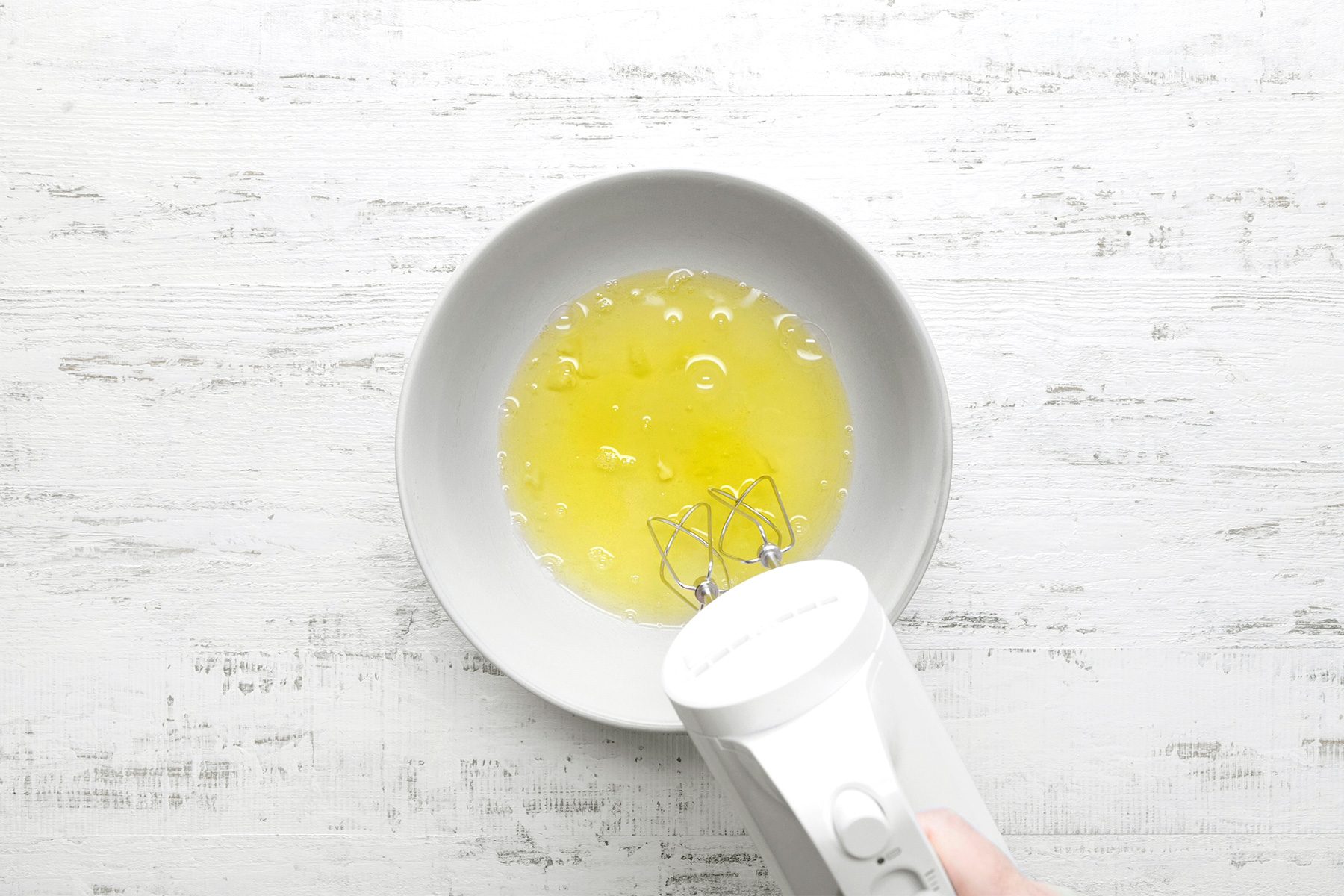 overhead shot of a white bowl contains egg whites, An electric mixer is being used to beat the egg whites into stiff peaks, The egg whites are a pale yellow color and have a frothy texture, The bowl is placed on a white wooden surface;