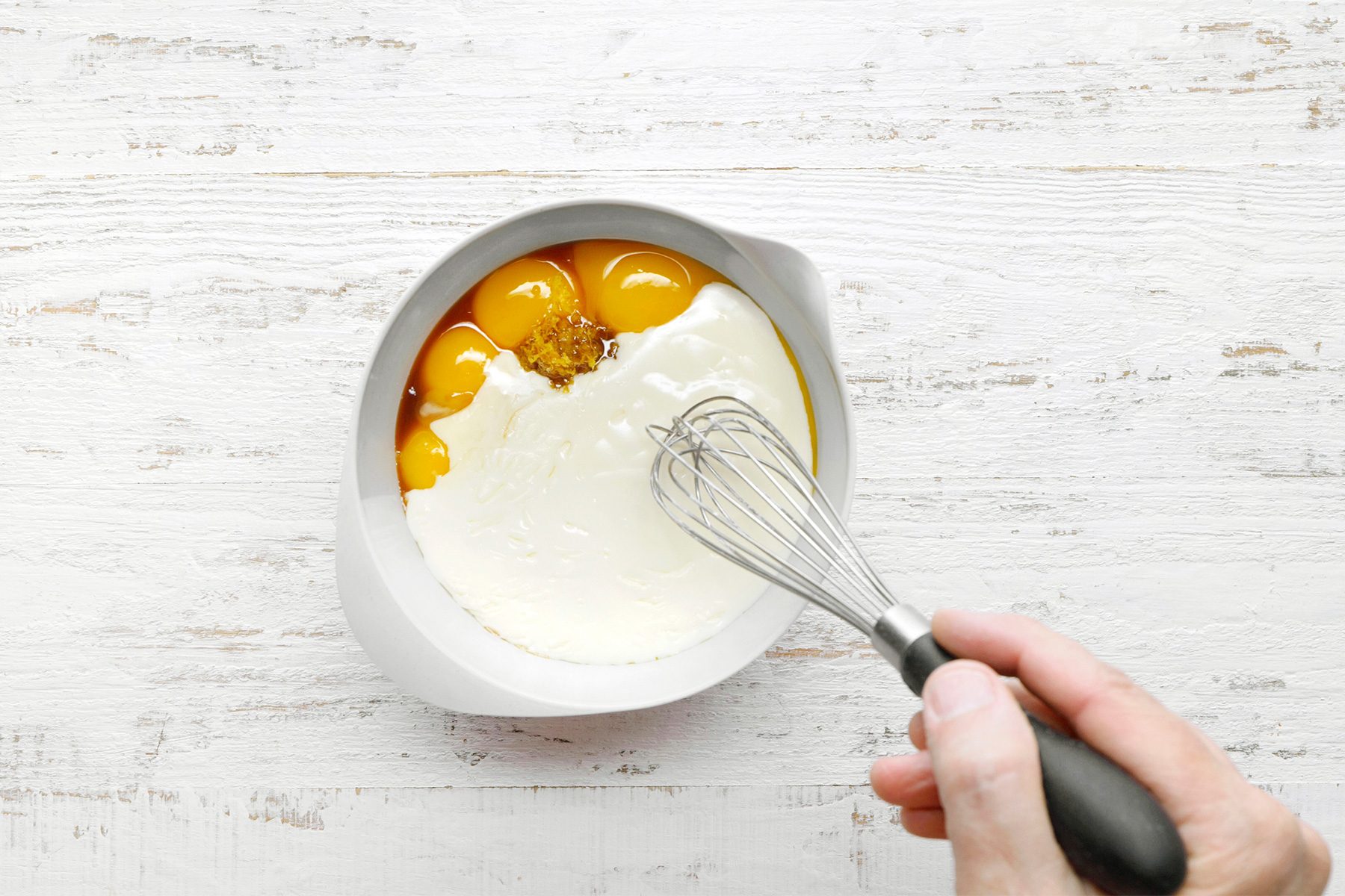 overhead shot of A white bowl contains a mixture of egg yolks, sour cream, and lemon zest, A whisk is being used to stir the ingredients together, The egg yolks are a vibrant yellow color, the sour cream is white, and the lemon zest is a light green color, The bowl is placed on a wooden surface;