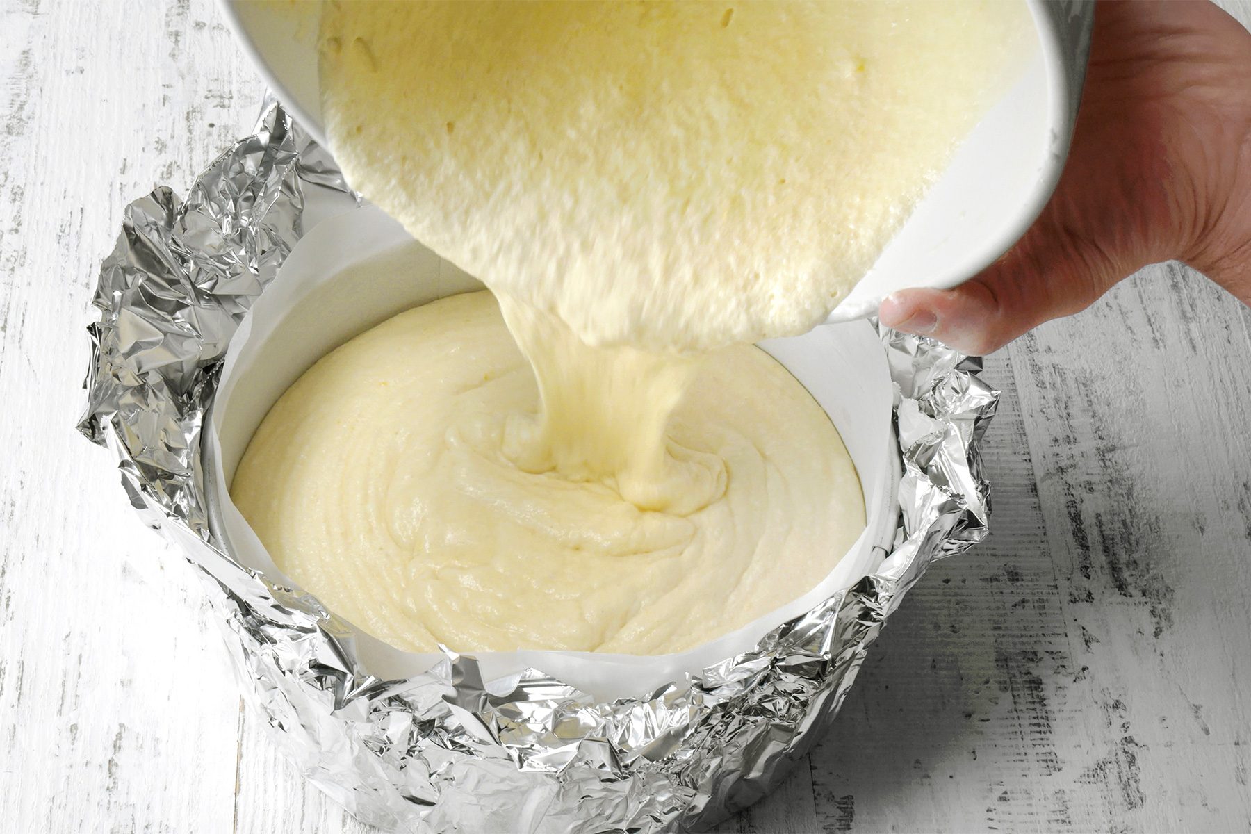 3/4th shot of a person pouring a creamy batter into a cheesecake pa, The batter is a pale yellow color and has a smooth texture, The cheesecake pan is wrapped in aluminum foil, and the batter is being poured evenly into the pan;