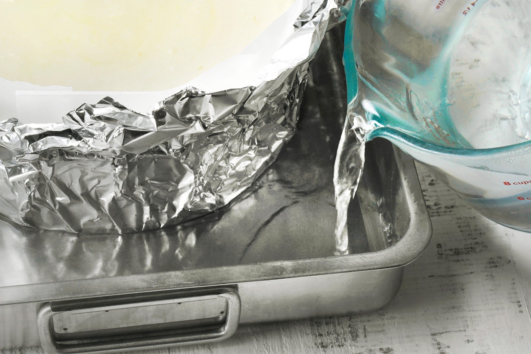 close shot of a baking pan wrapped in aluminum foil, A person is pouring water from a measuring cup into the baking pan, The water is creating a pool around the base of the pan, The image suggests that the baking pan is being prepared for a water bath;